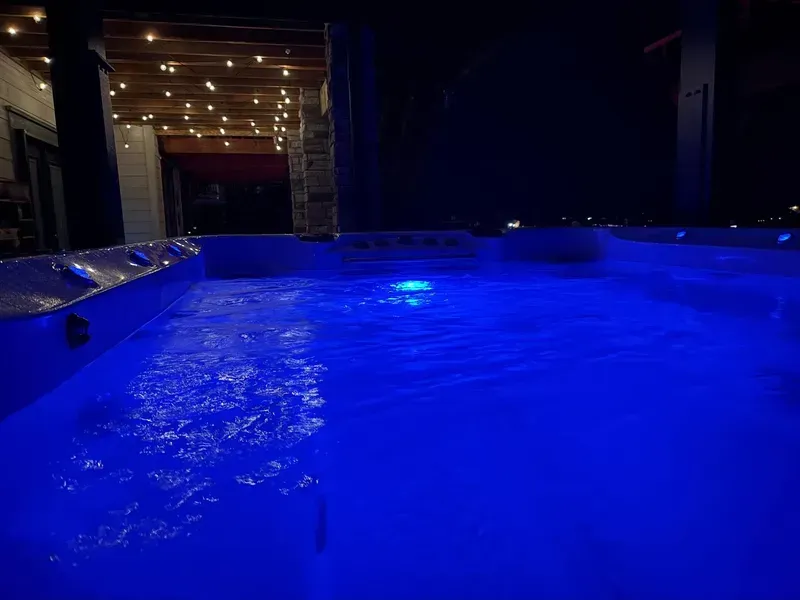 Blue-lit swimming pool at night. Water is rippling. String lights illuminate a nearby covered patio.