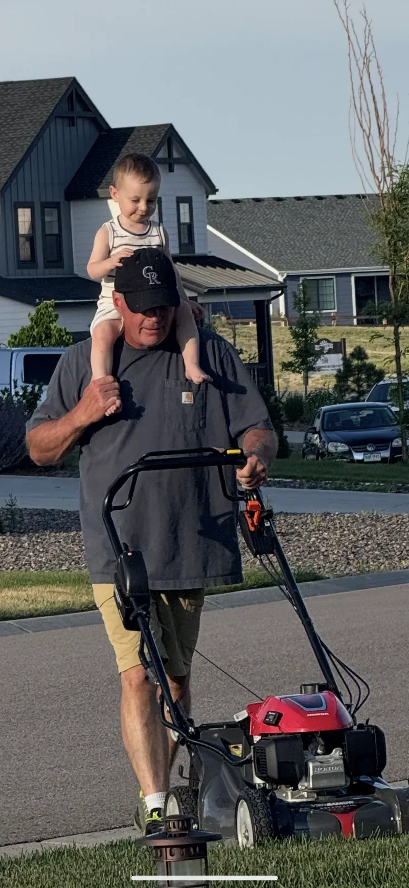 Man mows lawn with child on his shoulders.  Houses in background.