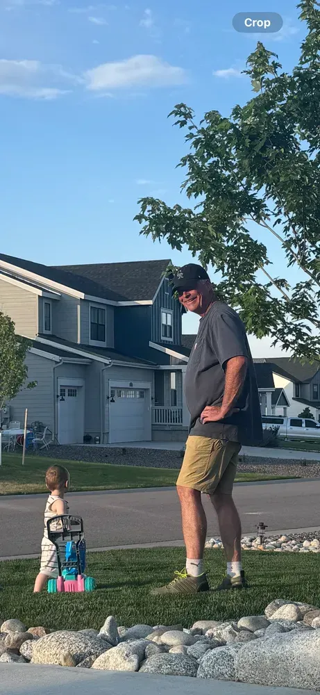 A man and a toddler stand on a lawn near a house. The toddler uses a toy walker.