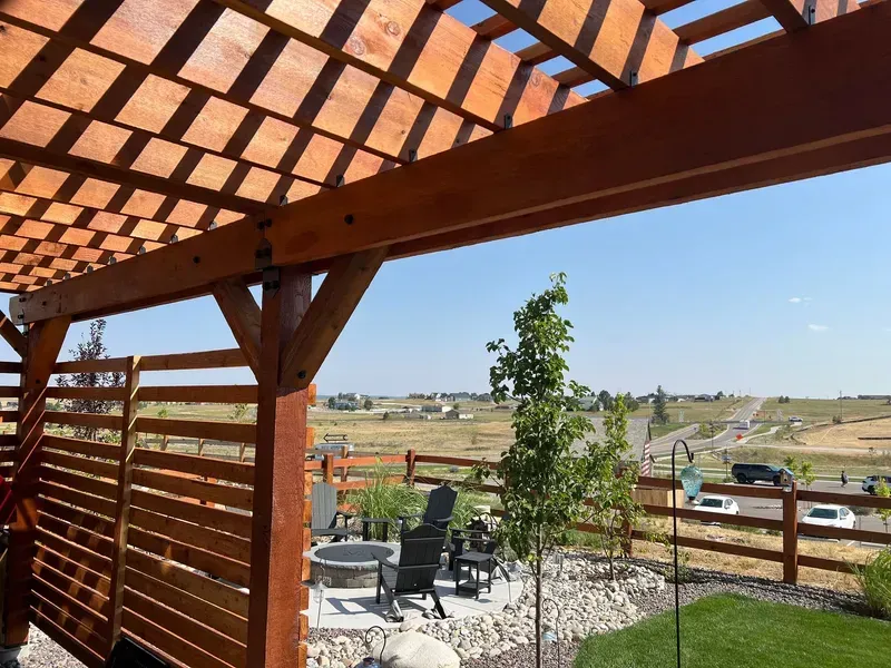 Wooden pergola with open lattice over a patio, overlooking a rural landscape on a sunny day.