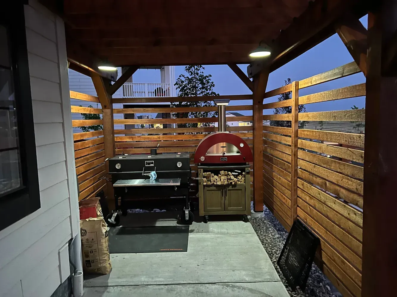 Outdoor cooking area with grill, pizza oven, and wood storage under a wooden pergola.