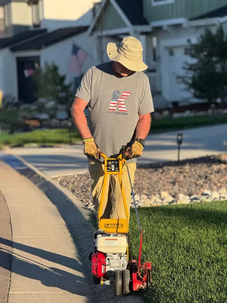 Man edging a lawn with a powered edger on a sidewalk, near houses with US flags.