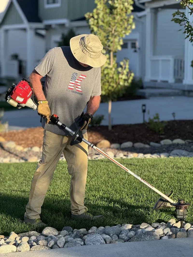 Man using a weed whacker near a sidewalk, trimming grass.