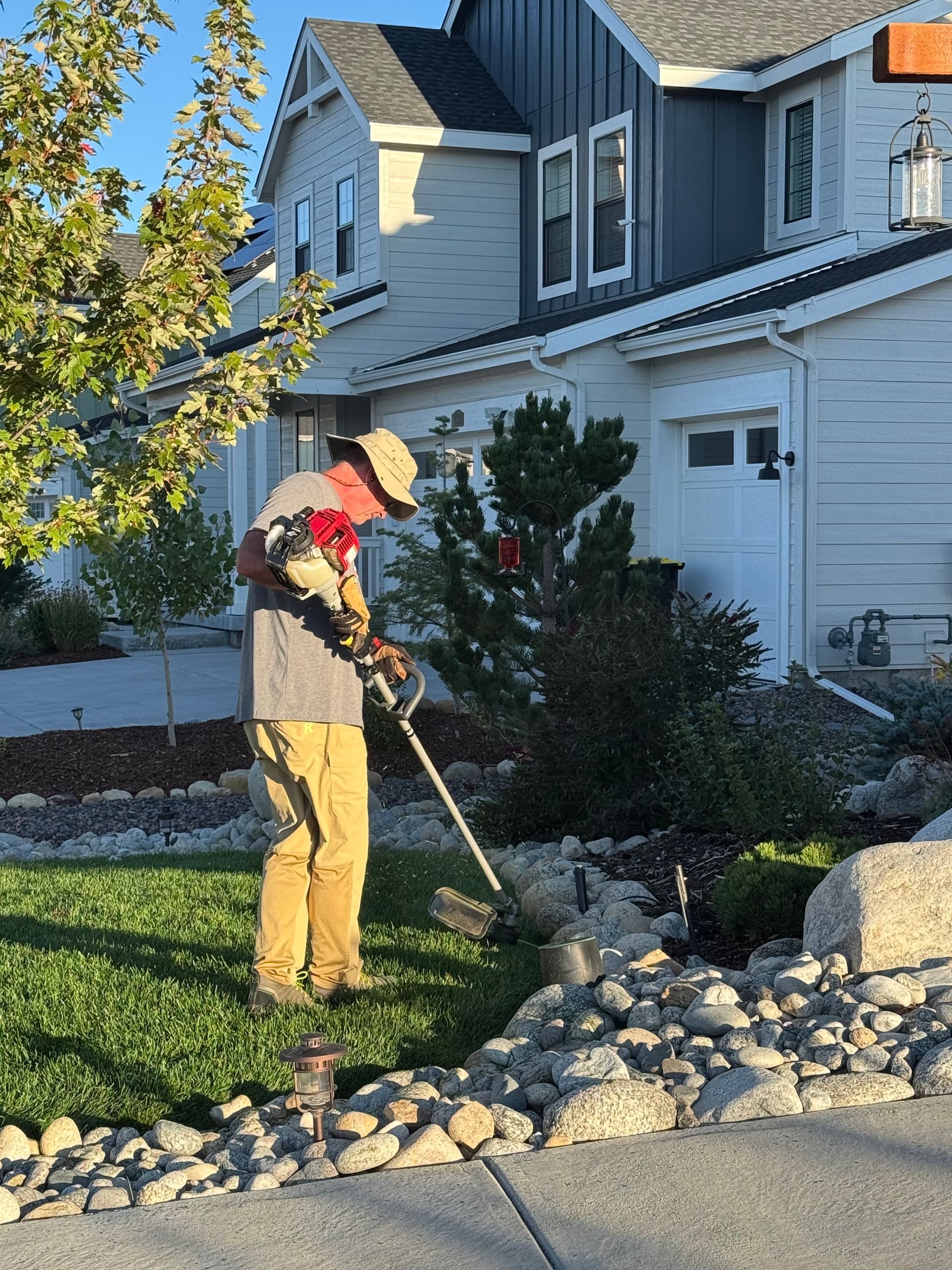 Person mowing a green lawn with a black and gray lawnmower in a sunny backyard.
