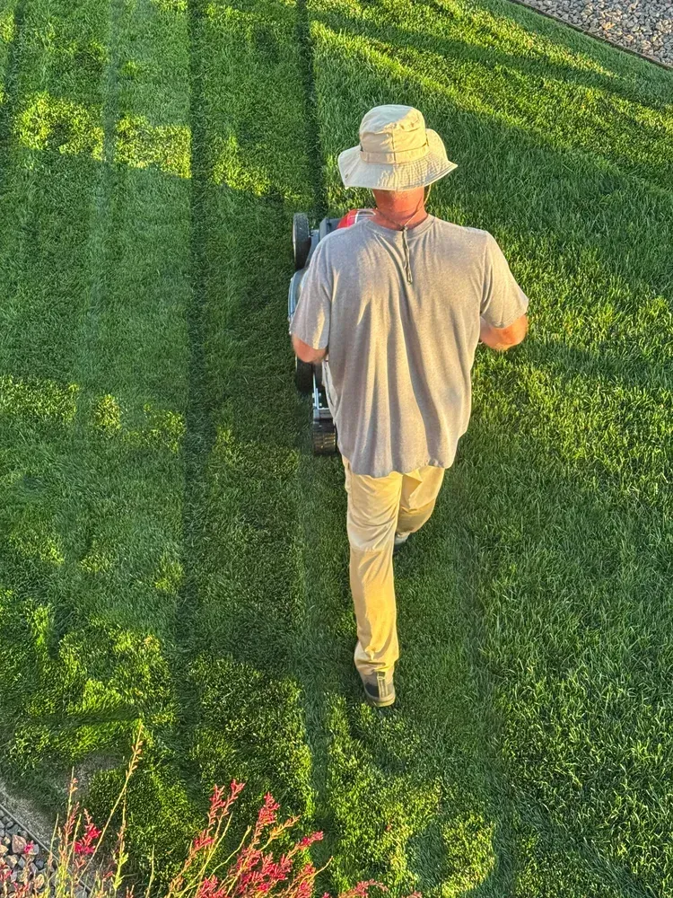 Man mowing a green lawn in a yard, wearing a hat, khaki pants and gray shirt.