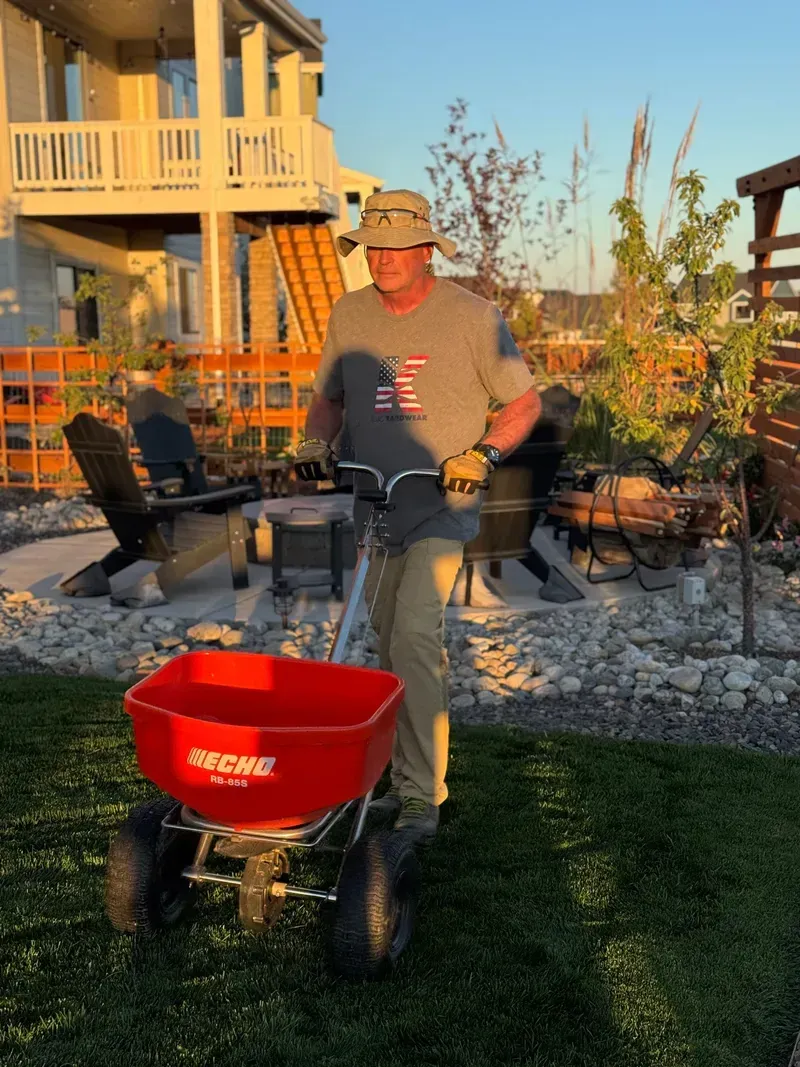 Man pushing a red ECHO spreader on a lawn, near a house and seating area.
