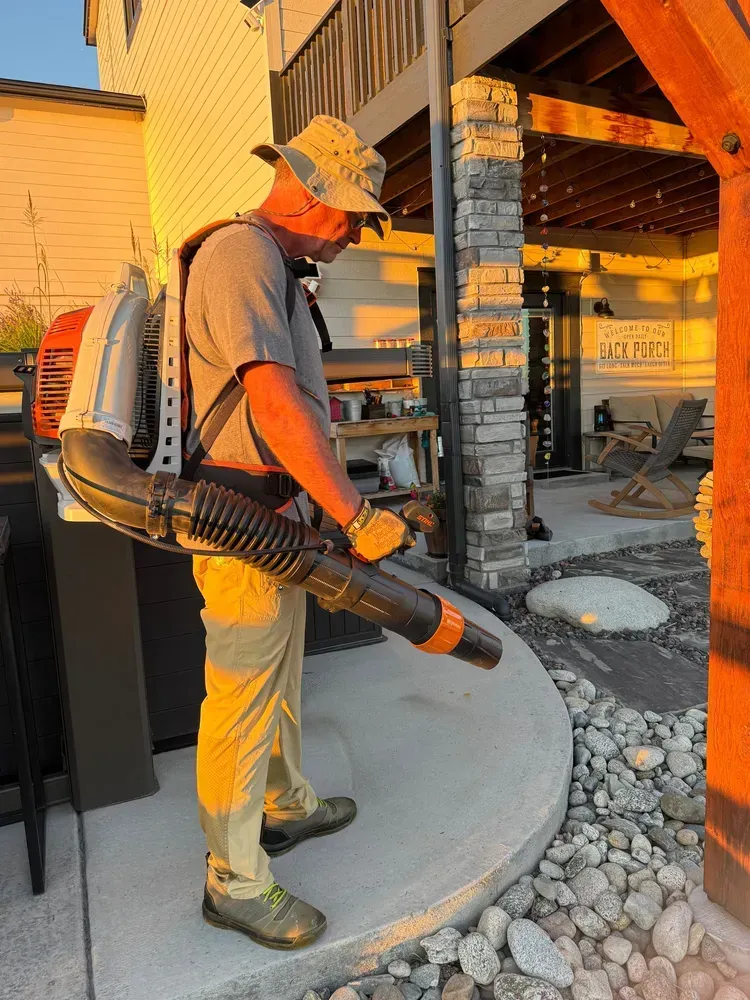 Man in a hat using a backpack leaf blower on a concrete patio, rocks and house in background.