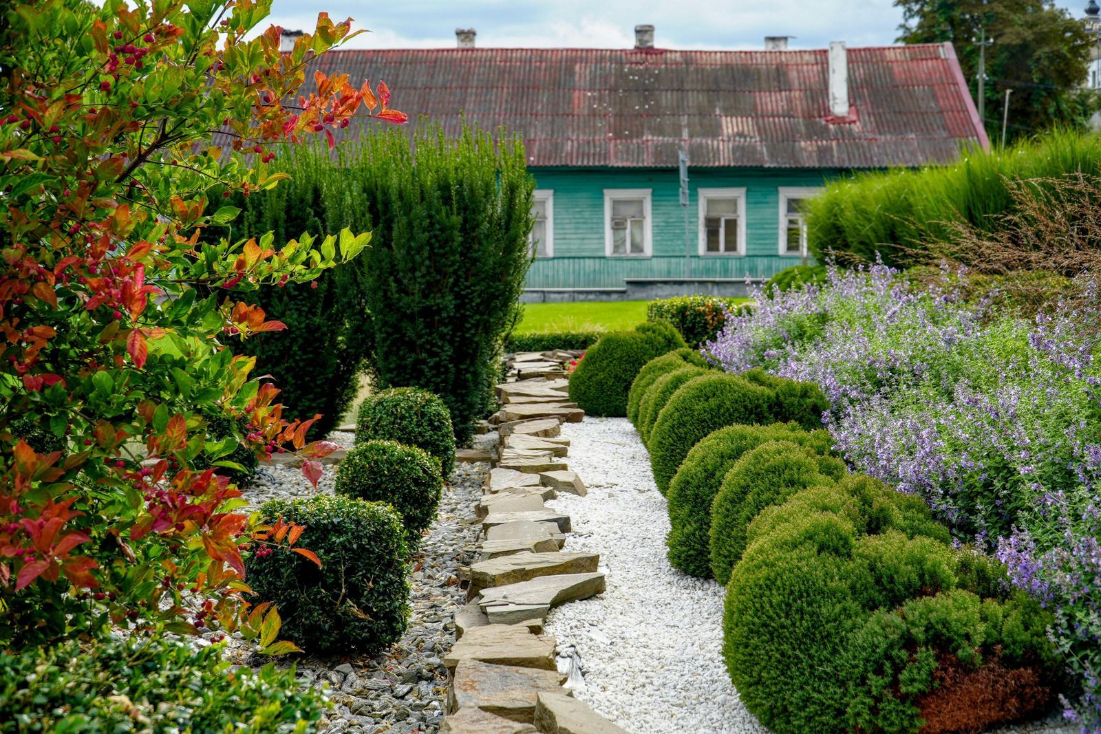 Stone path through a garden with green bushes, white stones, and a teal building in the background.