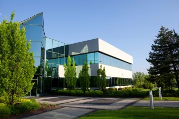 Modern office building with glass facade, surrounded by trees and a green lawn under a blue sky.