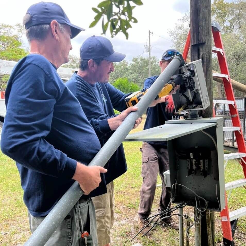 Three men are working on a pole with a ladder in the background.
