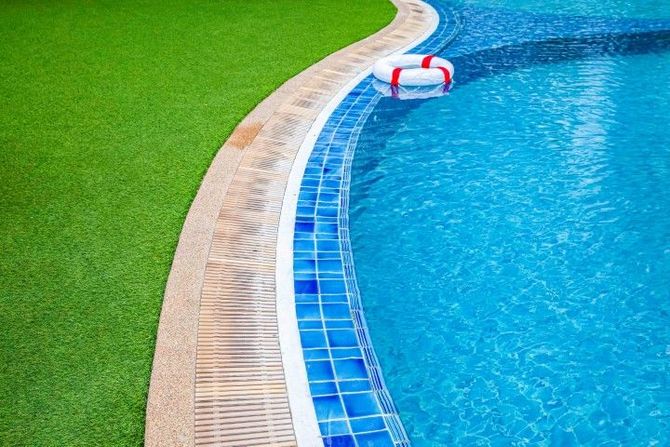 A lifebuoy floats in a blue tiled swimming pool next to a curved edge and a green grass lawn.