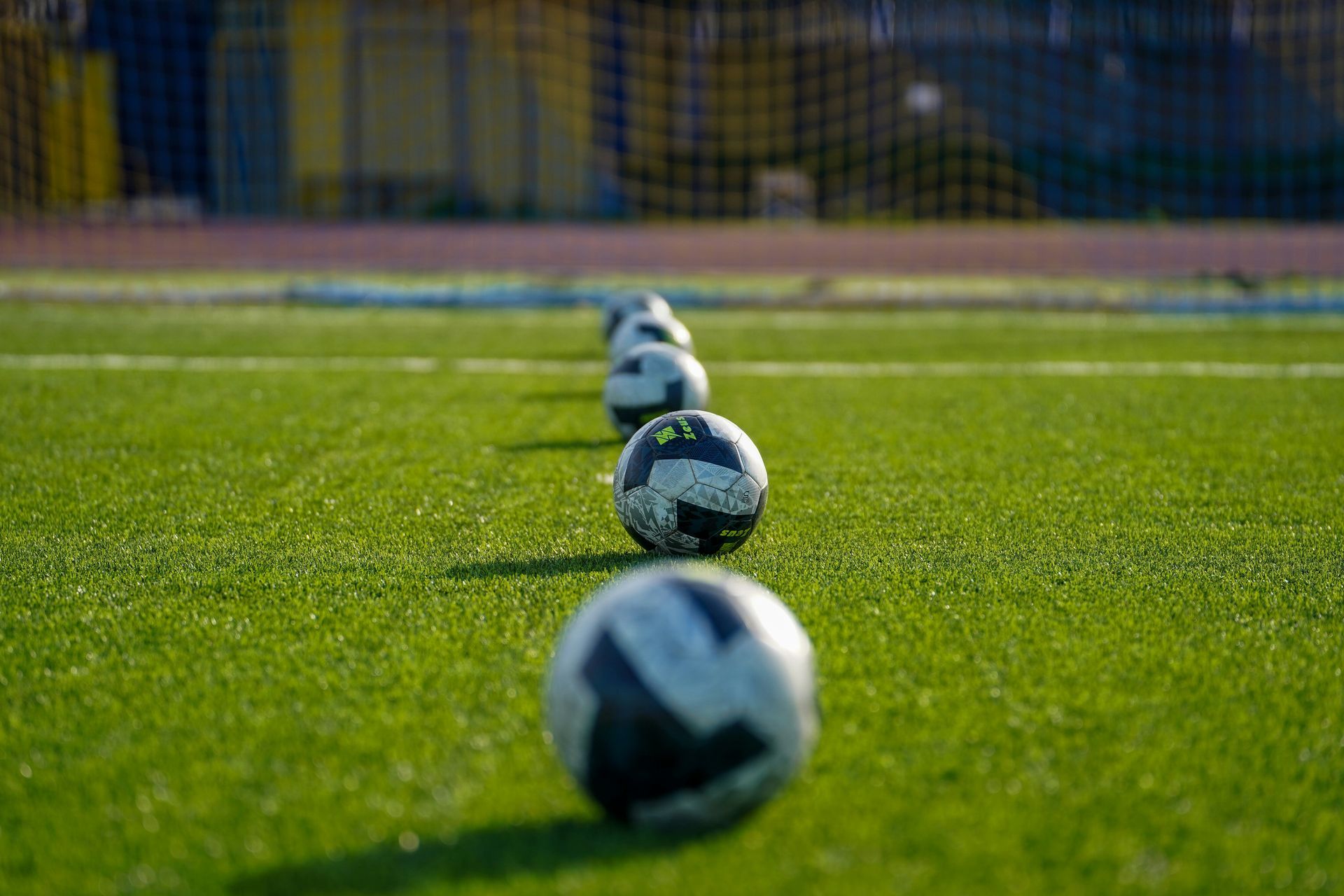 A line of soccer balls rests on a green turf field, with a goal net visible in the blurred background.