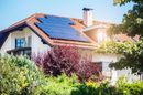 A house with solar panels on the roof is surrounded by trees and bushes.