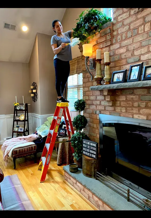 Amaya's Cleaning Service - A woman is standing on a ladder in a living room decorating a fireplace.