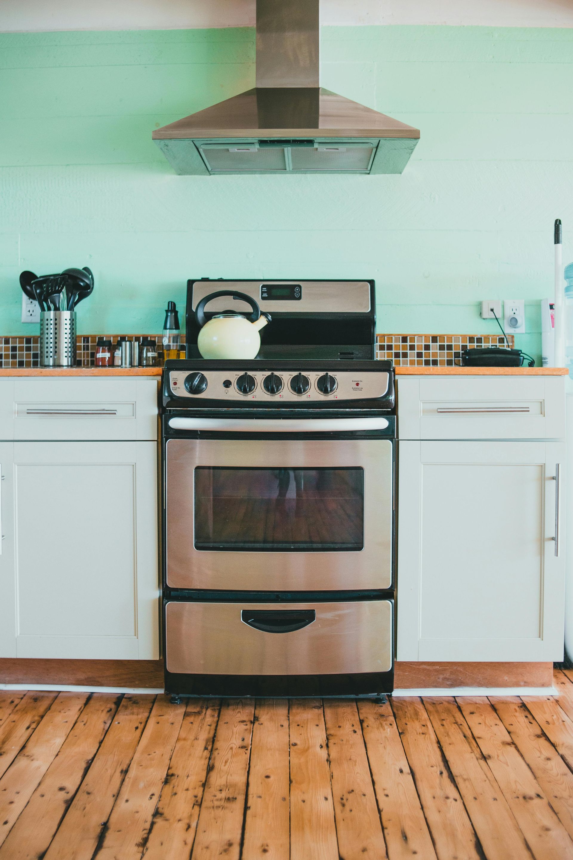 A stainless steel stove with a light yellow kettle sits between white cabinets against a mint green wall.