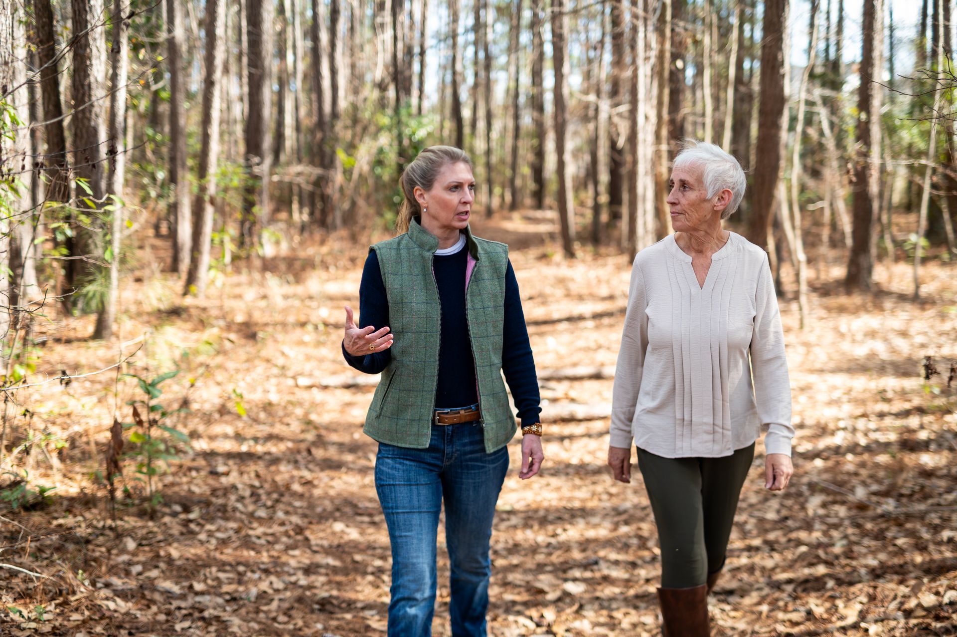 Two women walking and talking in a sunny forest. One gestures while speaking.
