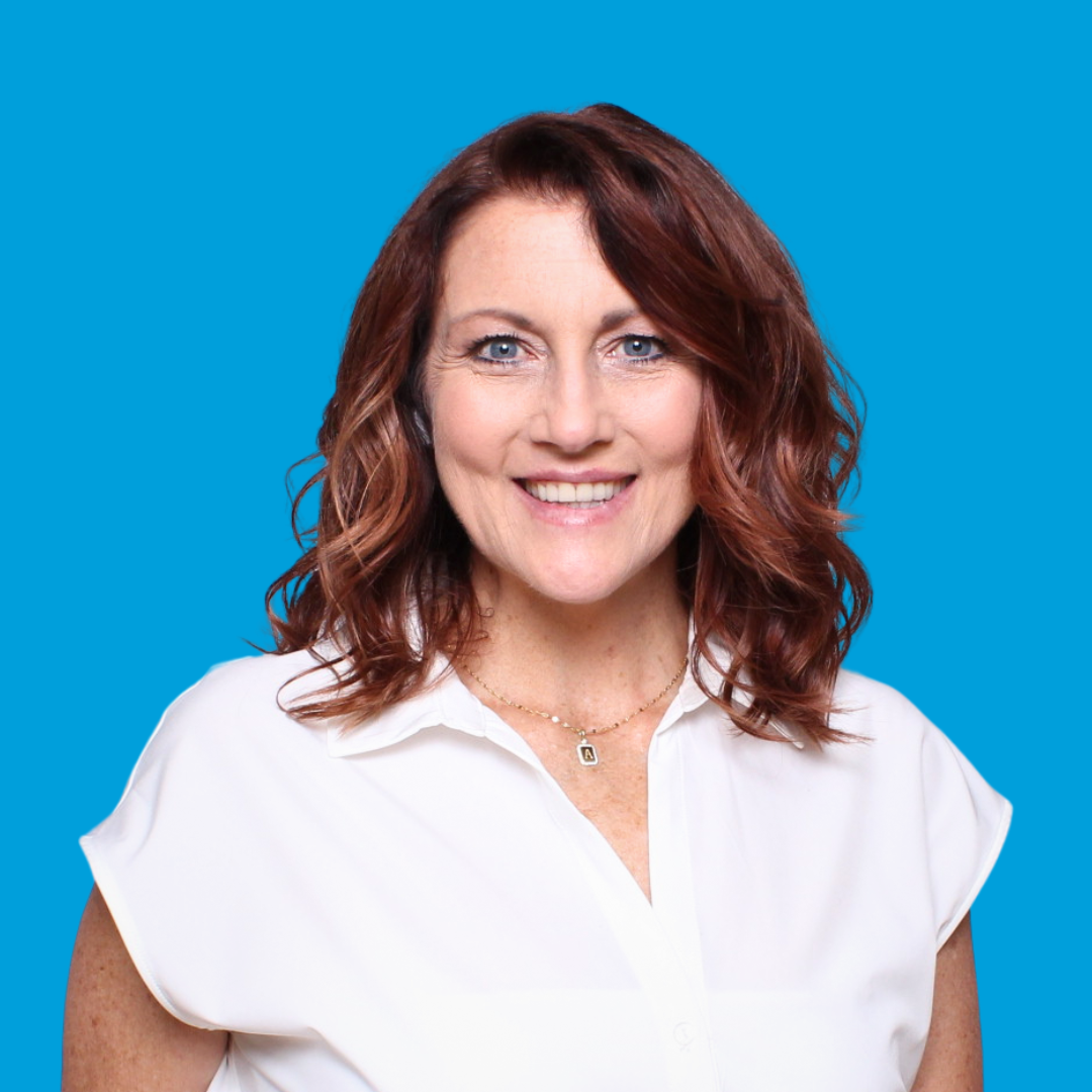 Woman with auburn hair smiles, wearing a white shirt against a blue background.