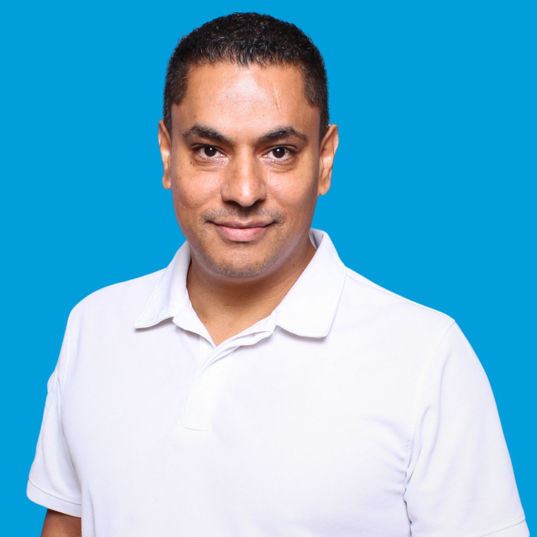 Man with short dark hair in white polo shirt, smiling, blue background.