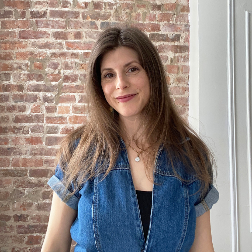 Woman with long brown hair smiles, wearing a denim shirt against a brick wall and white trim.
