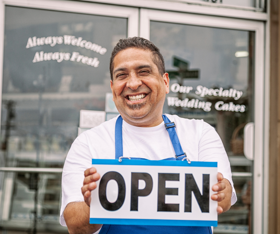 A man in an apron is holding an open sign in front of a store.