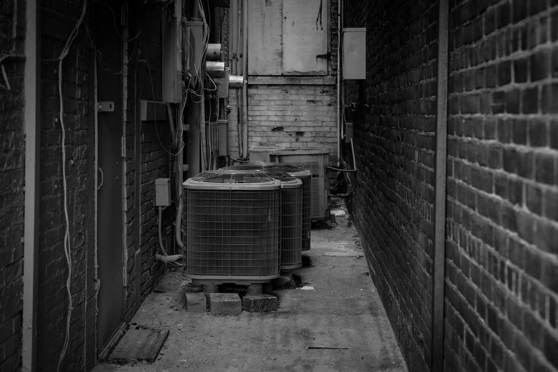 Narrow alley with brick walls, metal units, and a door at the end in black and white.