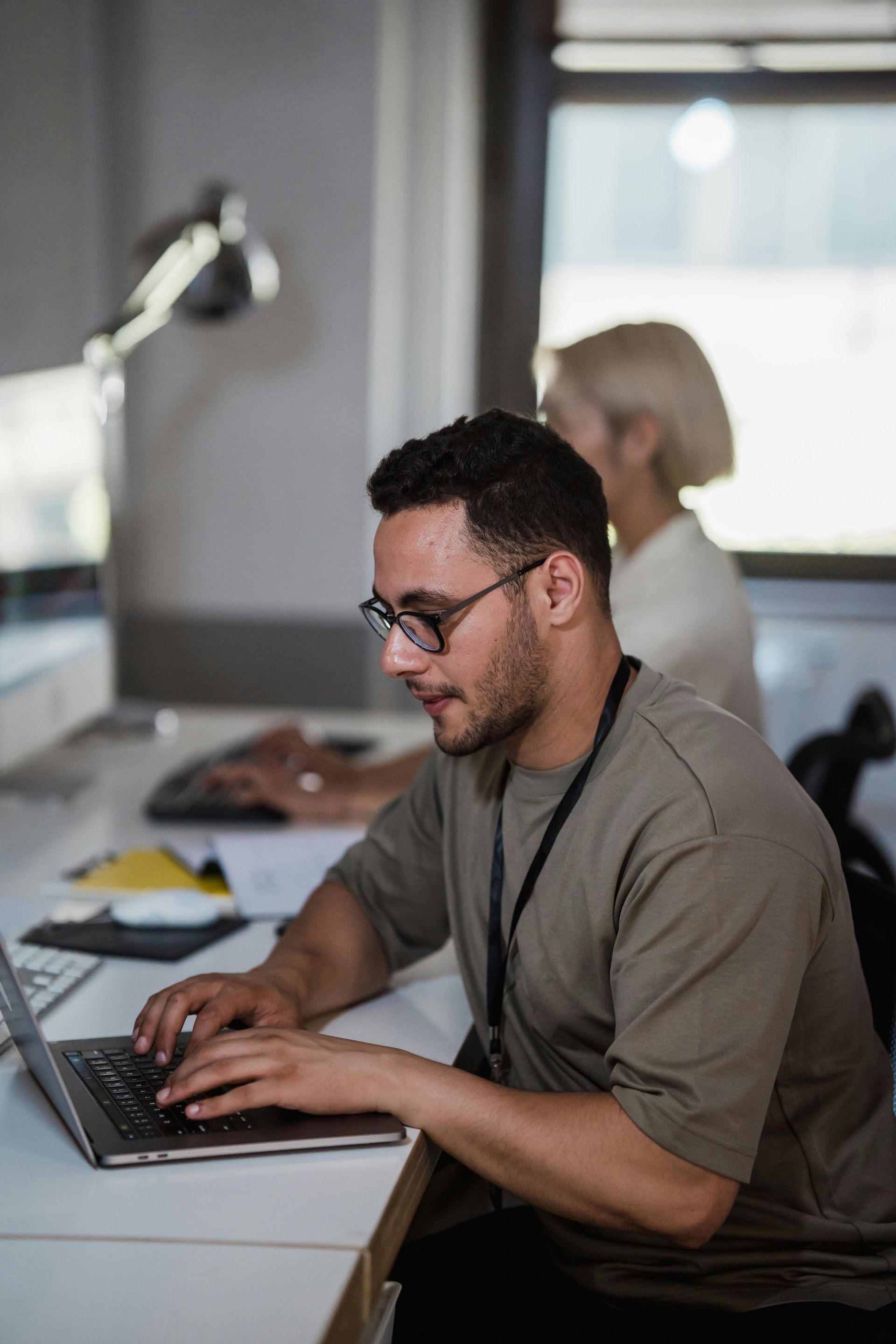 Person typing on a laptop at a desk in a bright office, with another person blurred in the background