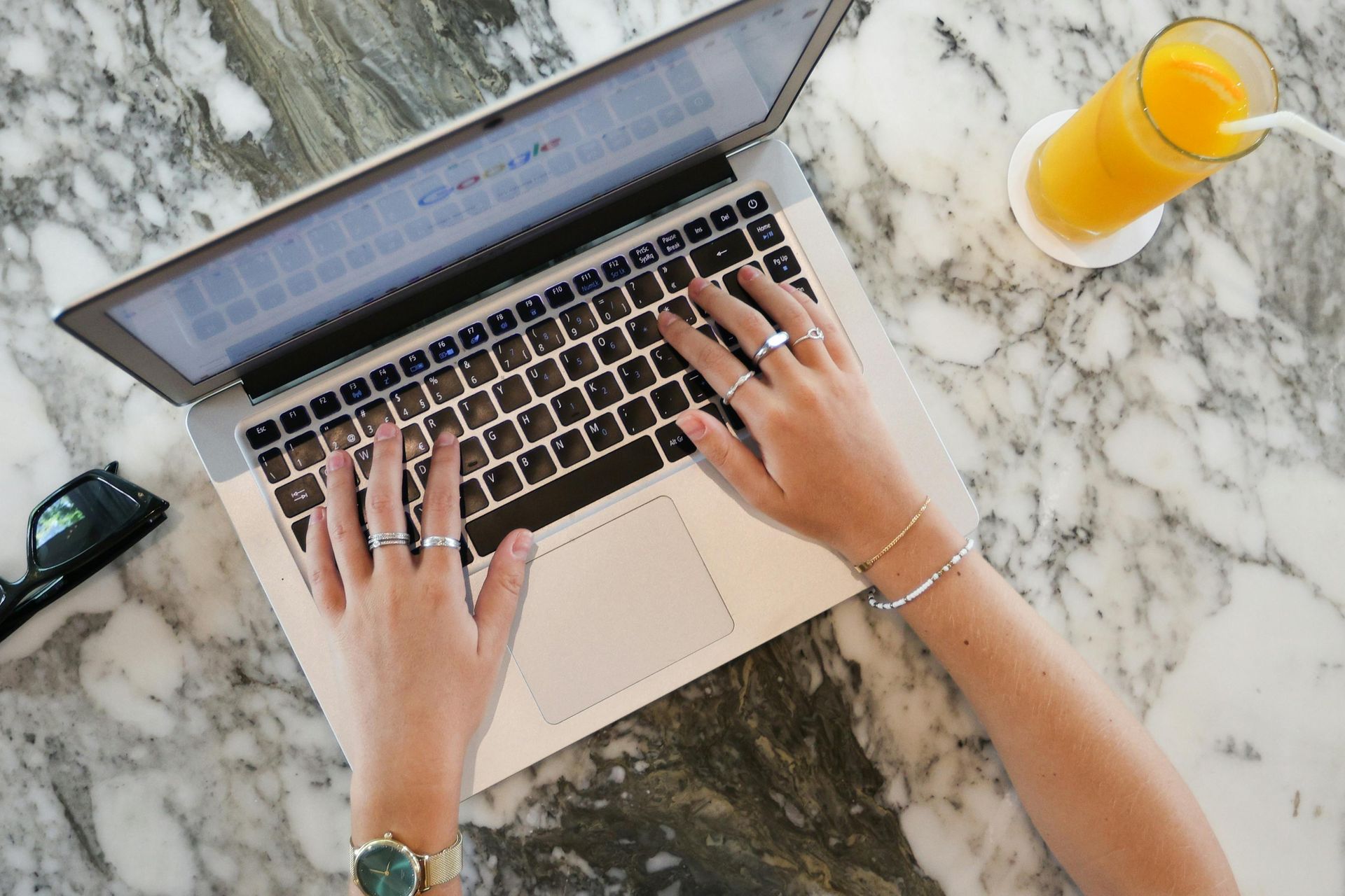 Hands typing on a laptop beside a glass of orange juice on a marble table
