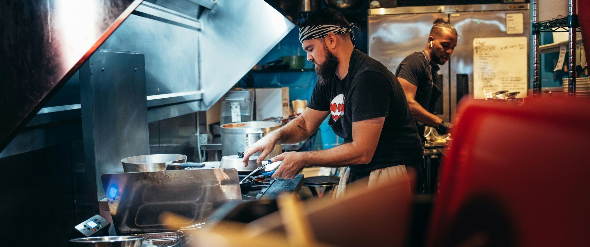Two cooks prepare food in a professional kitchen under an industrial exhaust hood.