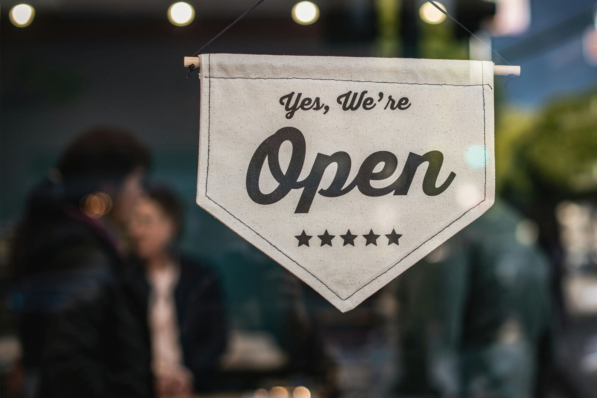 “Yes, we’re open” hanging sign in a shop window, with blurred people and lights in the background
