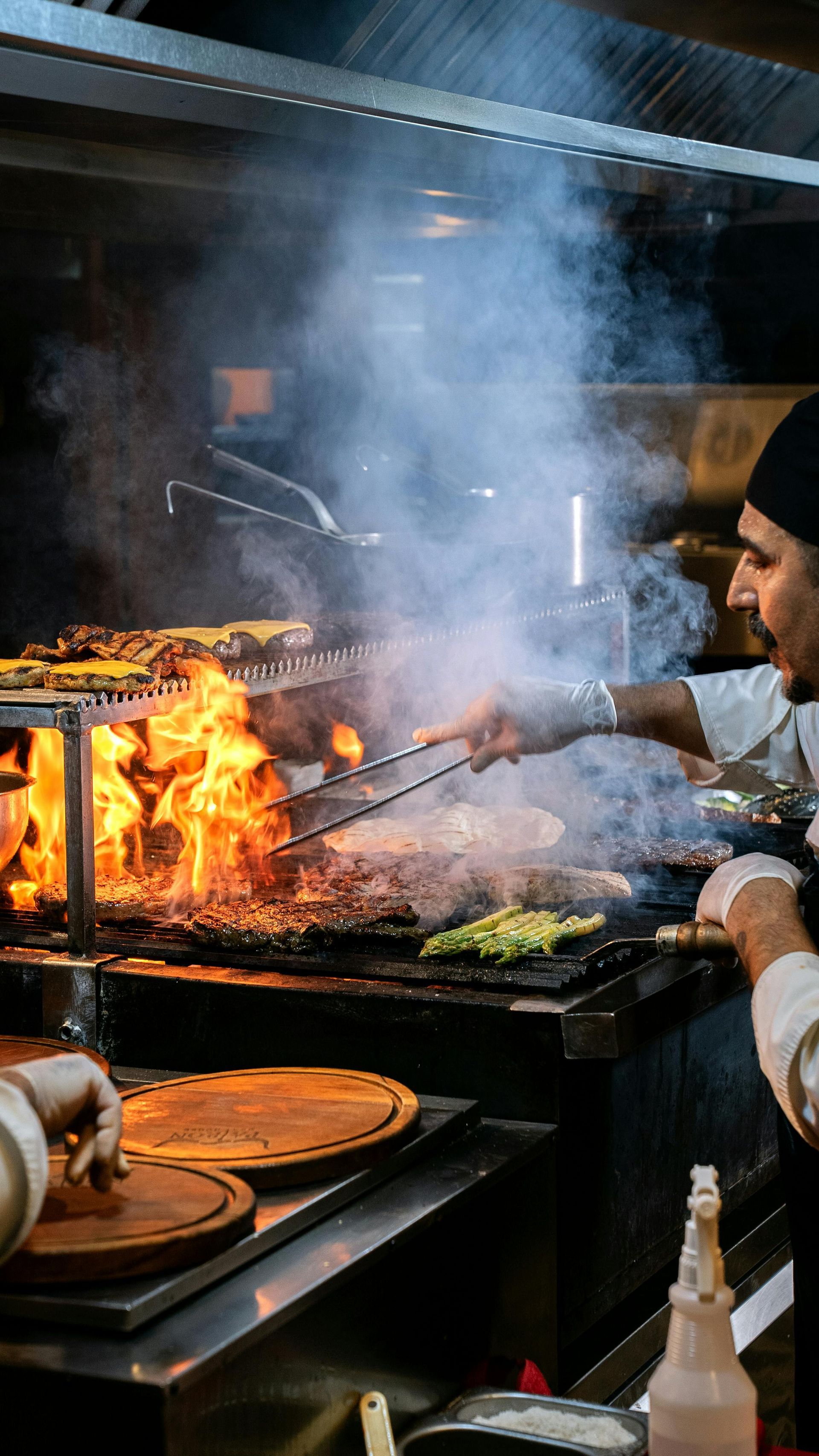 A cook in a black hat grilling meat over an open flame with visible smoke in a kitchen.