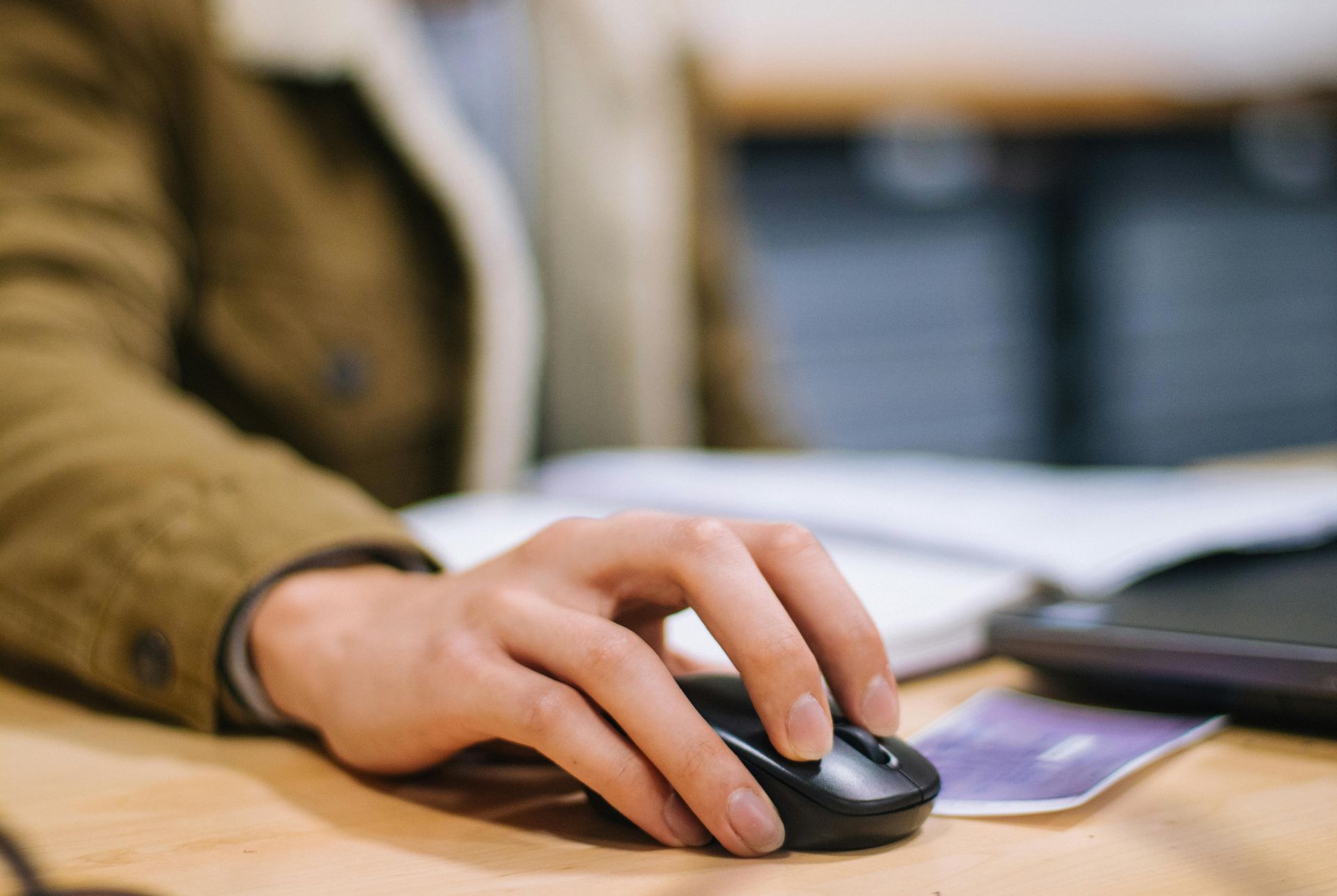 Hand using a black wireless mouse on a desk beside a blurred laptop and notebook