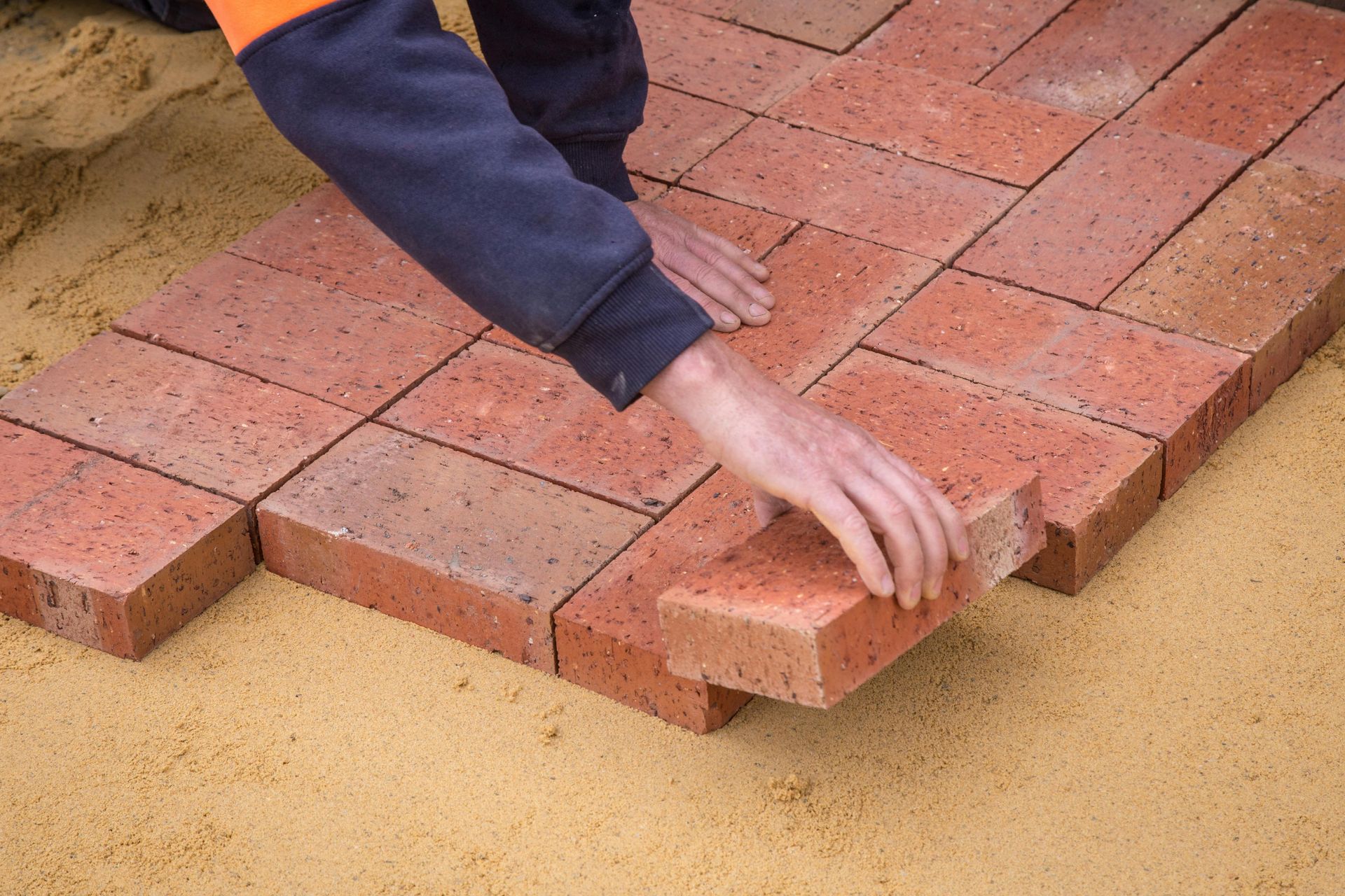 A person’s hands laying rectangular red bricks in a herringbone pattern on a sand base.