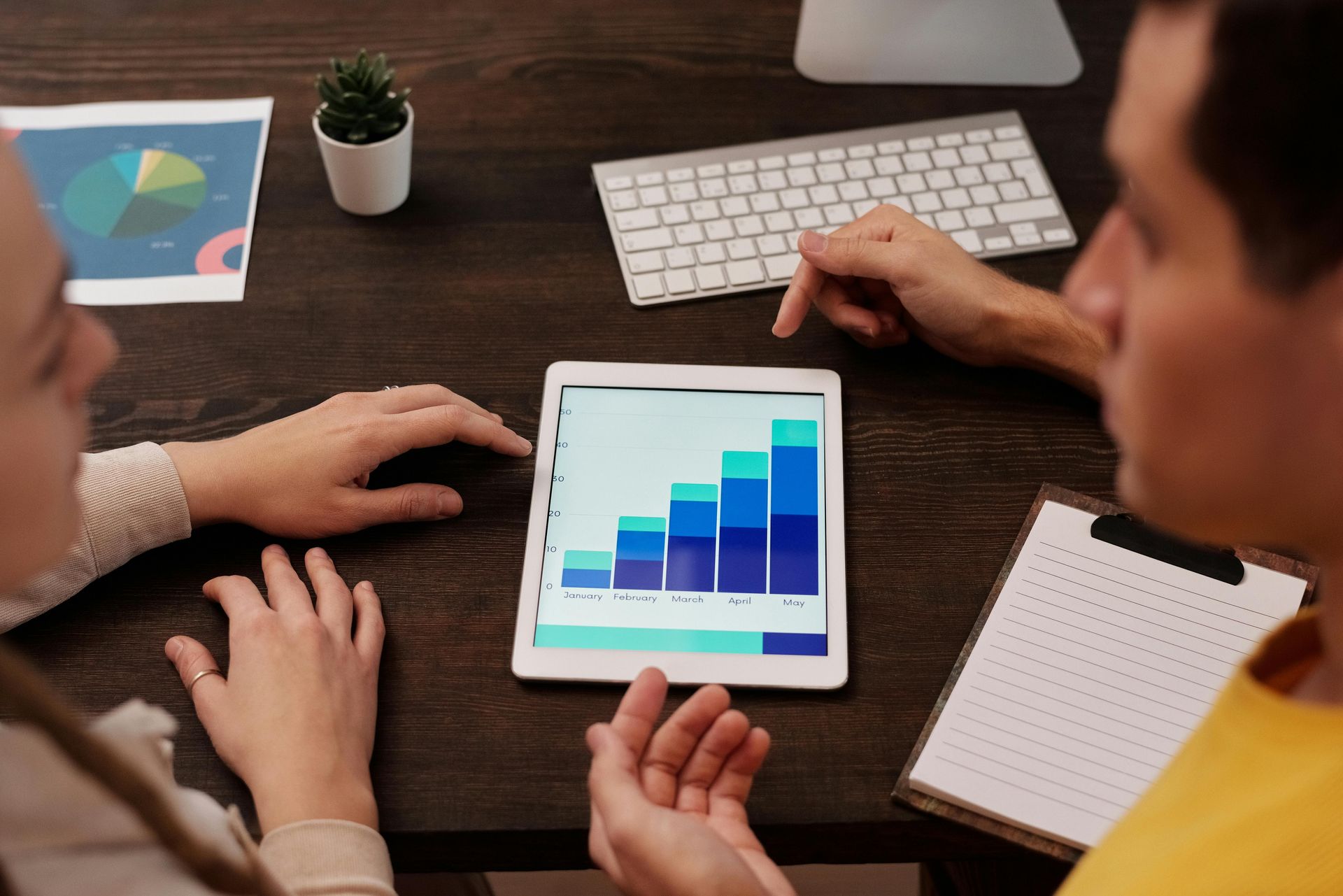 Two people review a tablet bar chart at a desk with laptop, keyboard, notebook, and coffee.
