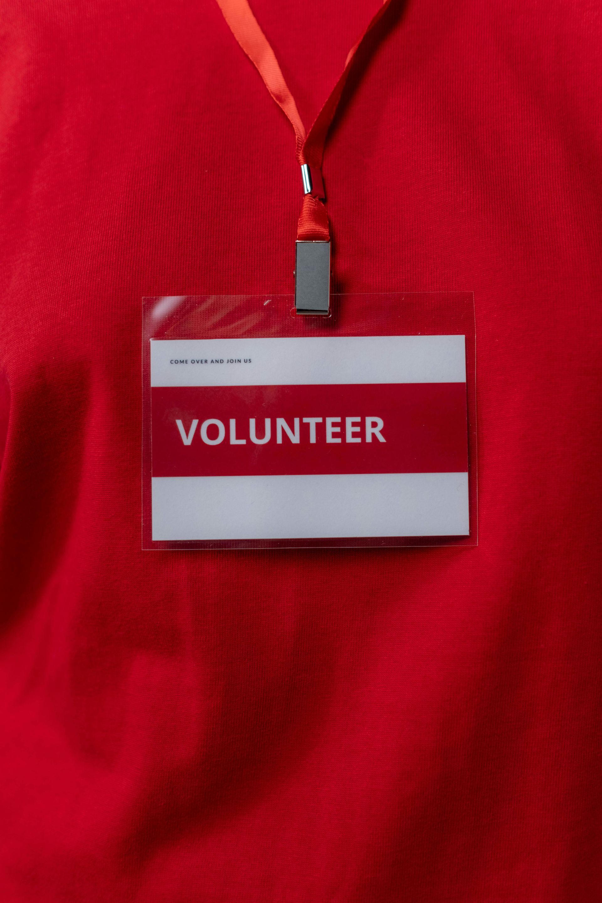 A volunteer name tag badge in a clear plastic holder hanging on a red shirt by a red lanyard.