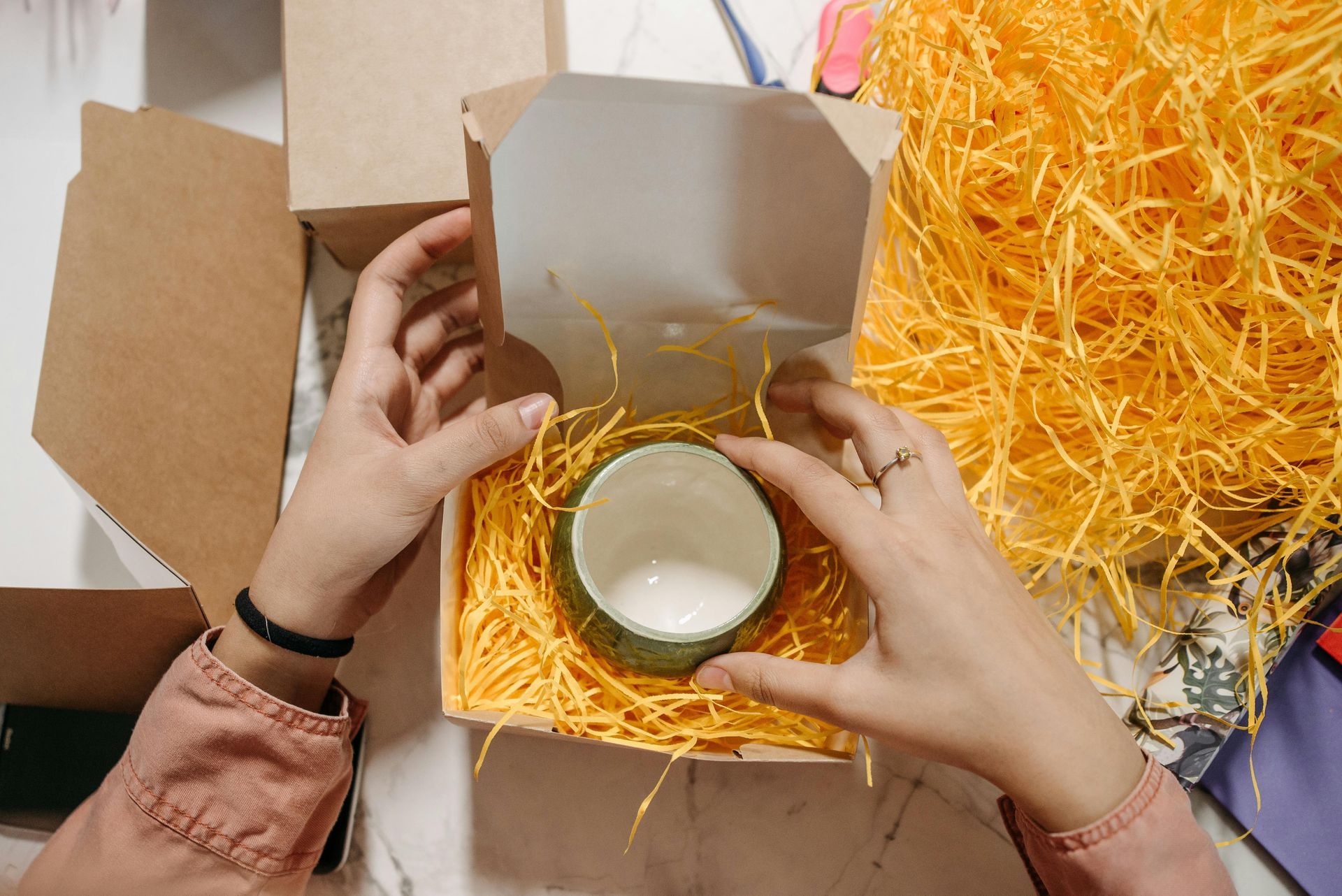 Hands place a small, green ceramic bowl into a gift box filled with yellow paper shred.