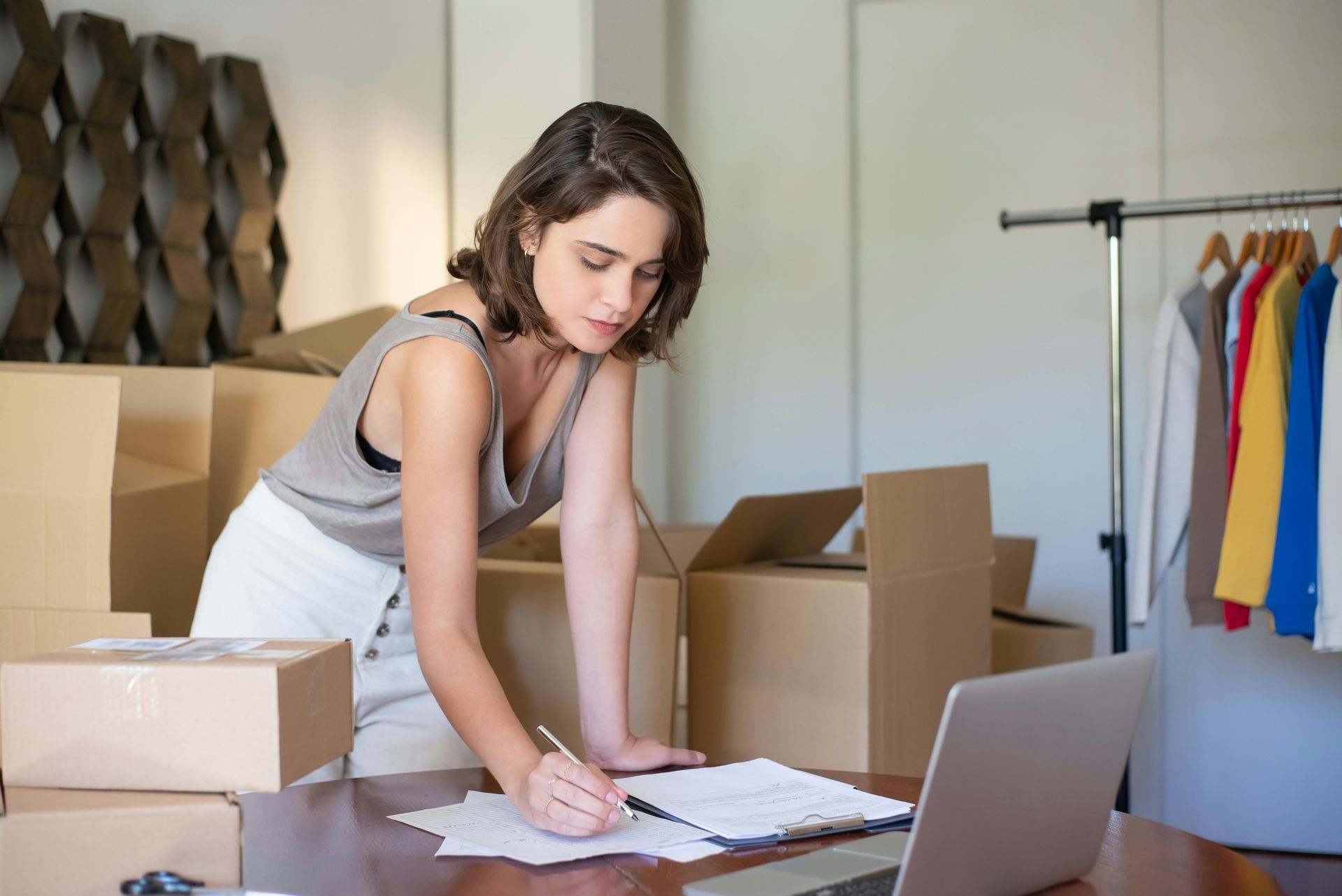Woman packing orders at a desk with boxes, paperwork, and a laptop in a small office.