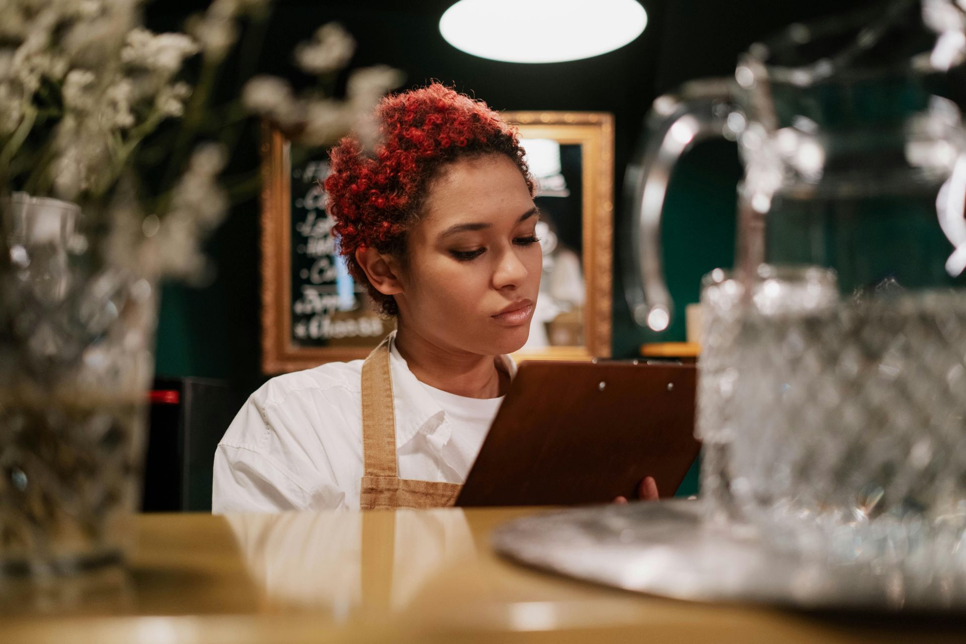A person with short, red hair wearing an apron focuses intently while writing on a clipboard in a dimly lit café setting.
