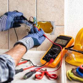 Electrician wiring an outlet with gloves, multimeter, tools, and hard hat on a workbench