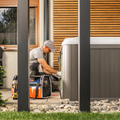 Worker kneeling beside a gray hot tub, repairing equipment in a modern backyard.