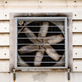 Industrial wall vent with large beige exhaust fan behind metal grille and dirty white siding.