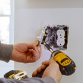 Hands installing an electrical outlet in a wall with a yellow power drill nearby