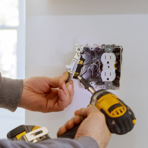 Hands installing an electrical outlet in a wall with a yellow power drill nearby