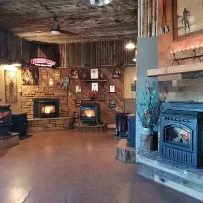 Cozy rustic room with wood-paneled walls, stone fireplace, and several wood stoves on display