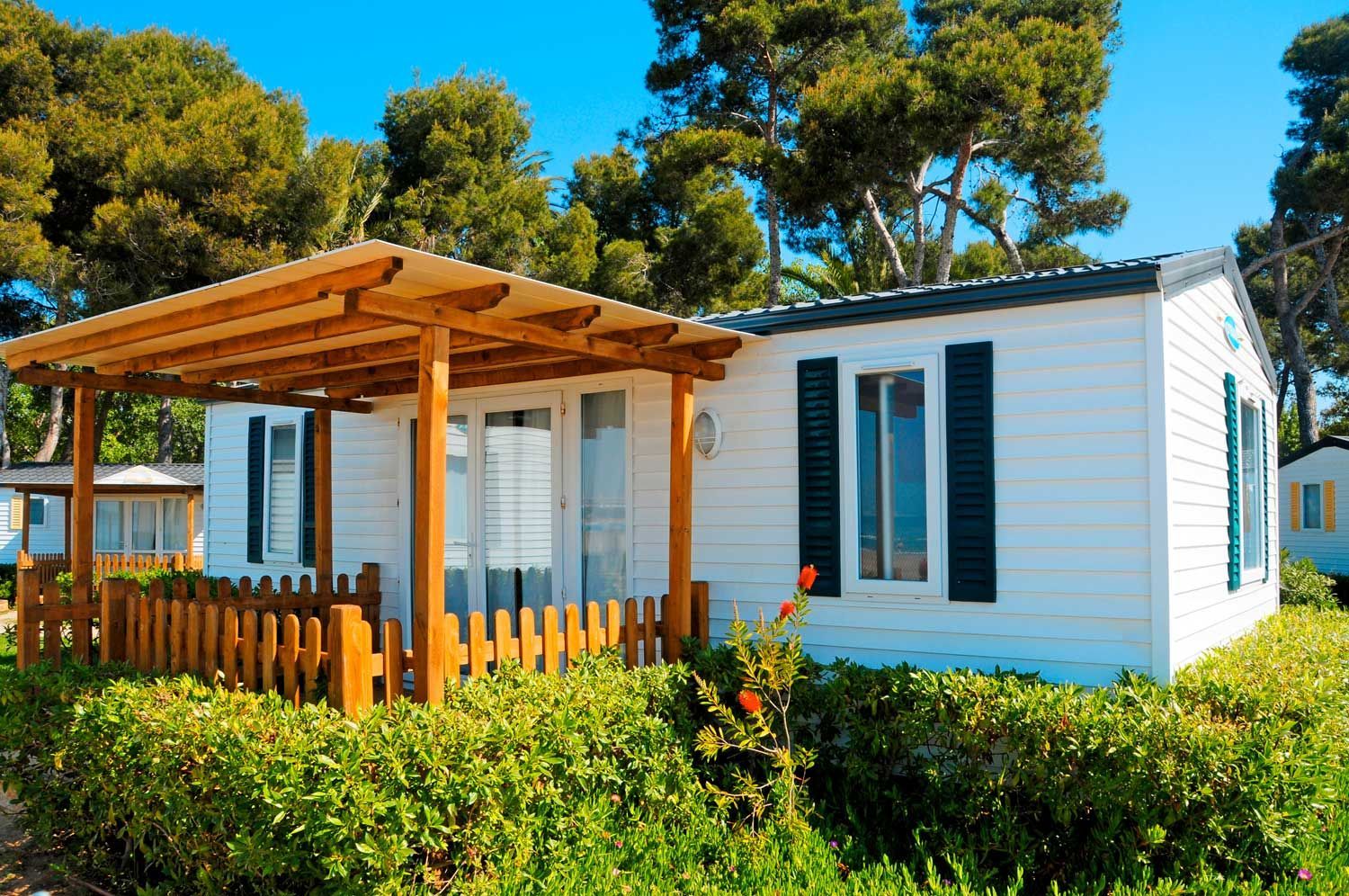 Mobile home with a wooden porch and green shutters surrounded by trees and shrubs.