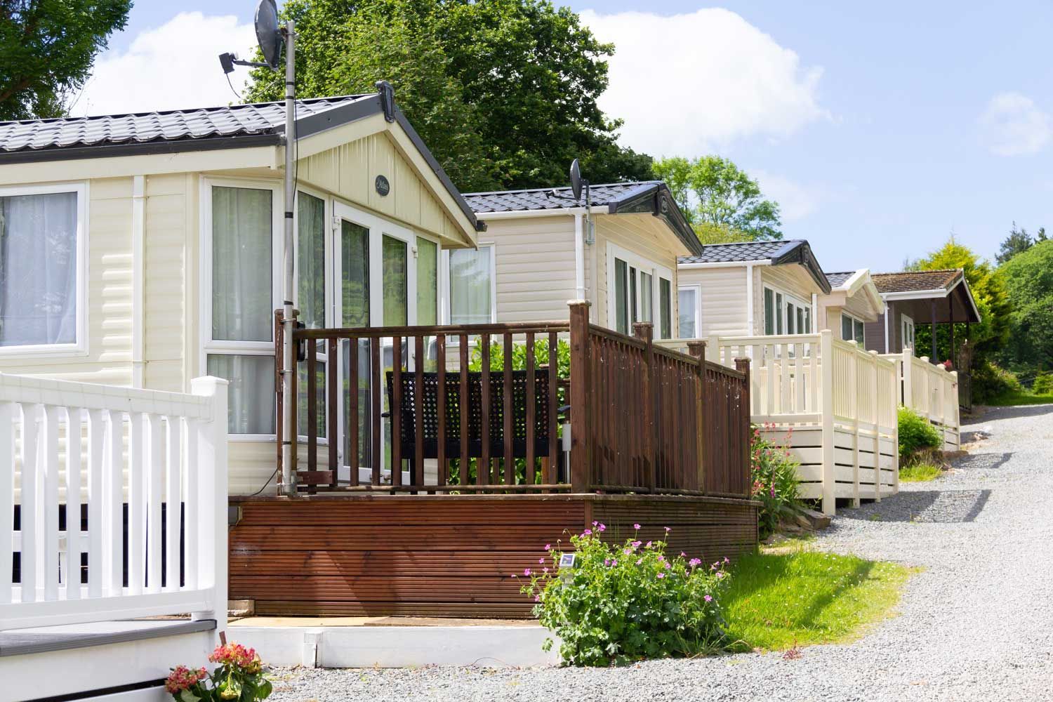 Line of parked mobile homes with raised decks along a gravel path surrounded by greenery.