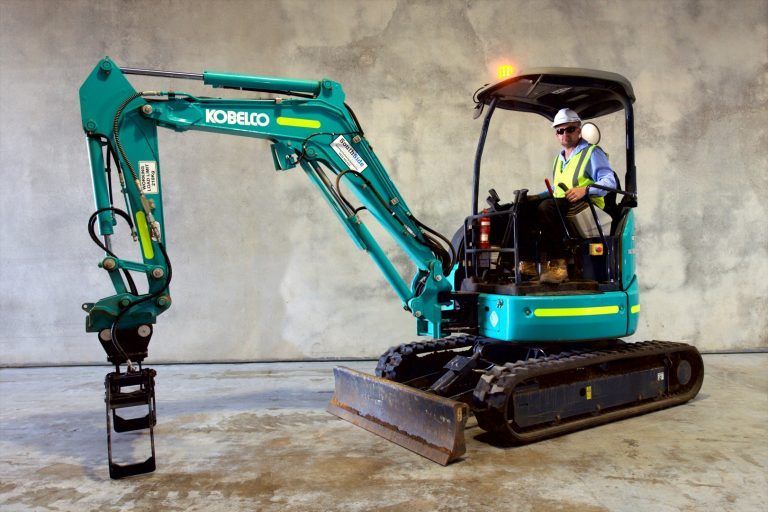A man is driving a kobelco excavator on a concrete surface.