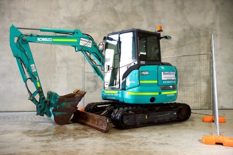 A small blue excavator is parked in a garage next to a fence.