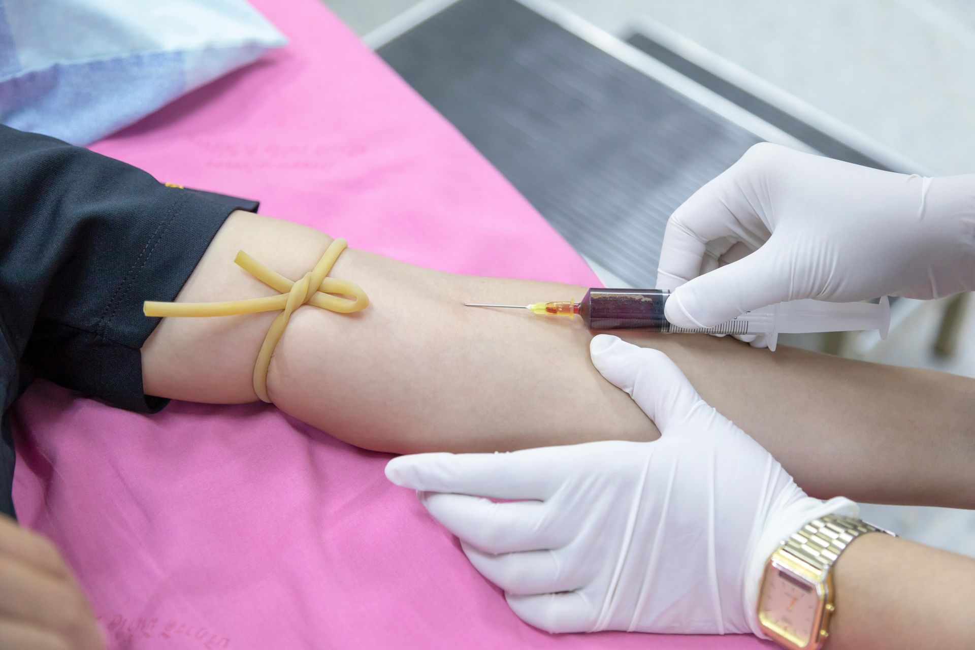 Medical professional drawing blood from a patient's arm. Arm has a tourniquet, gloved hands, setting appears to be a medical facility.