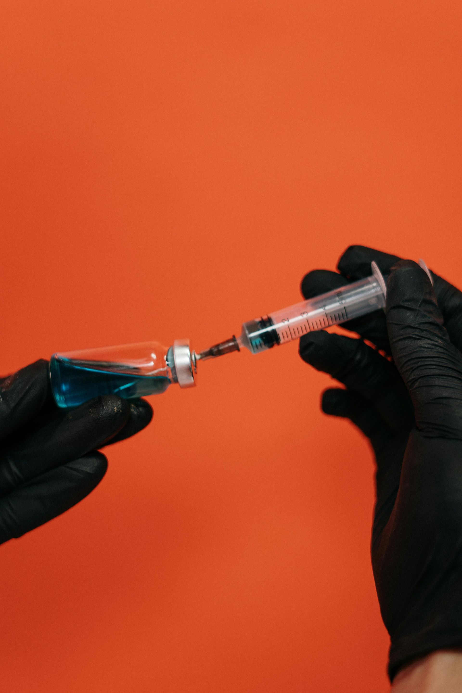 Hands in black gloves fill a syringe from a vial of blue liquid, against an orange background.
