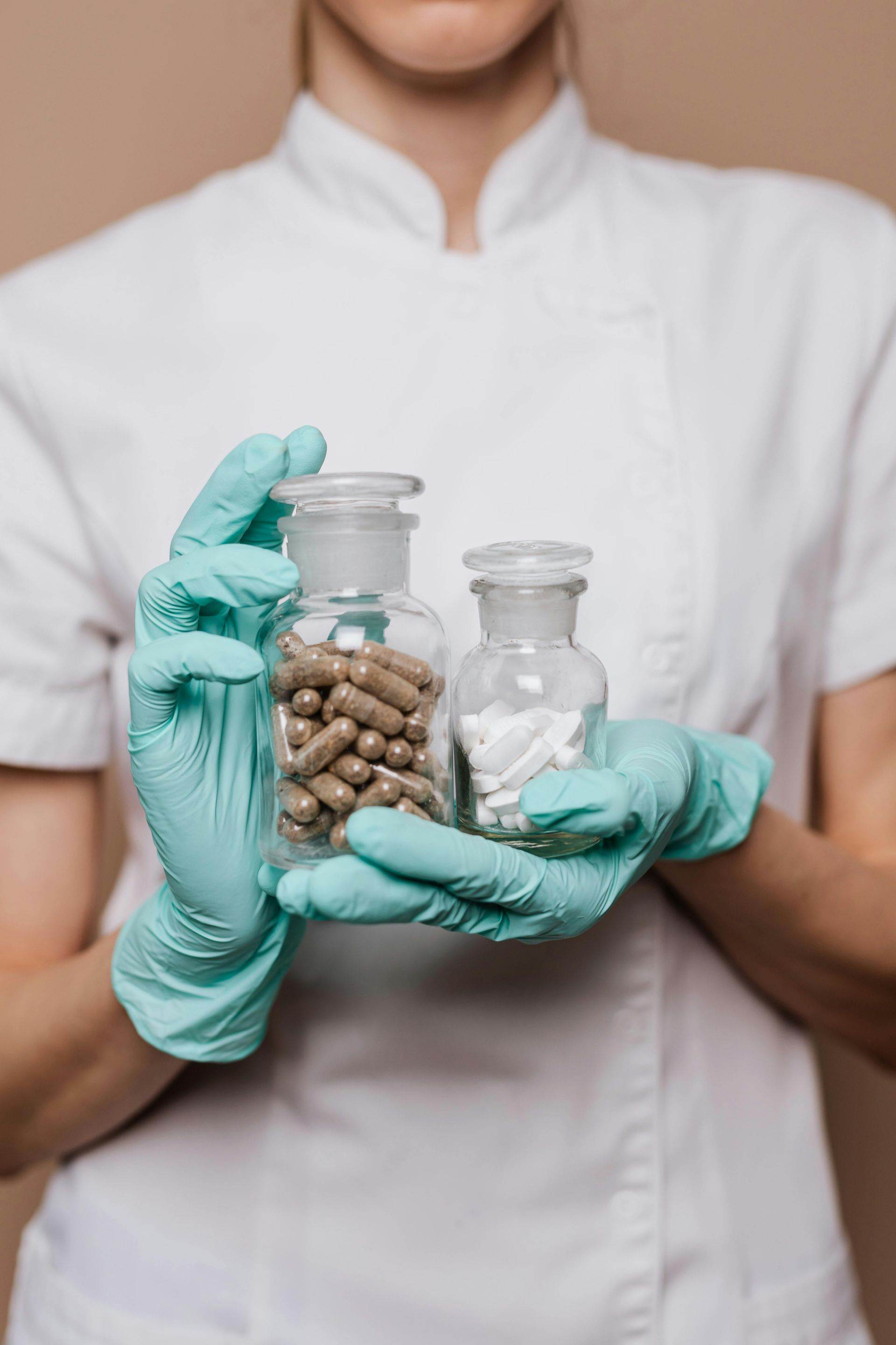 Person in white coat and gloves holding two glass jars of pills.